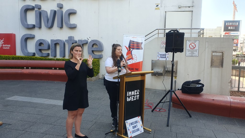 A white woman with long hair stands behind a podium saying Inner West Include Don't exclude and smiles into a microphone. 