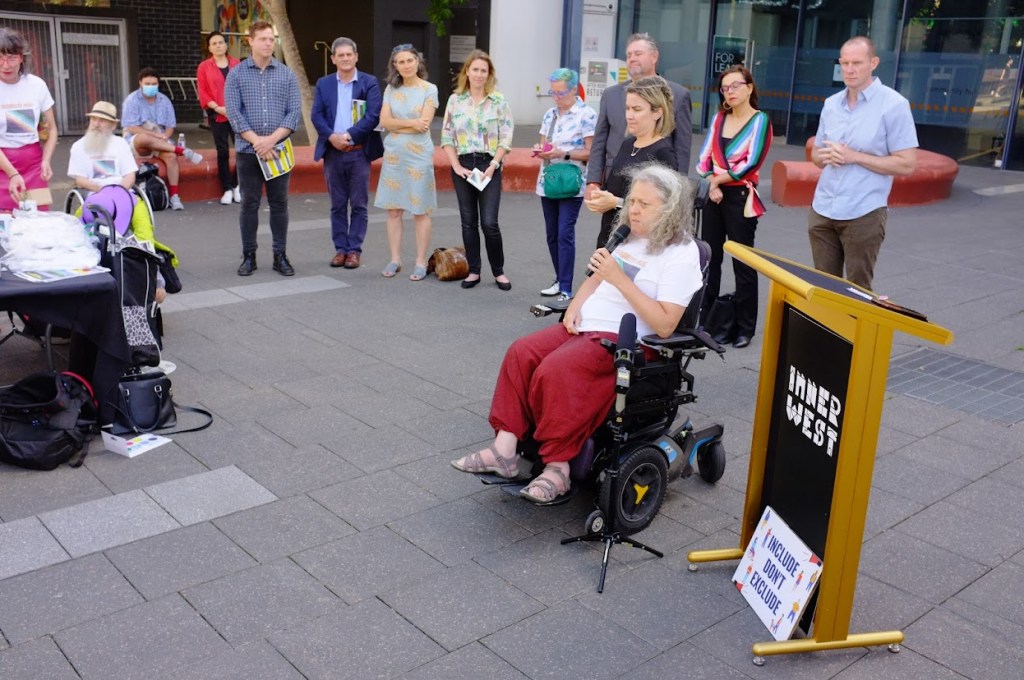 A woman in a power wheelchair with long curly hair holding a microphone. An Auslan interpreter is behind her. 